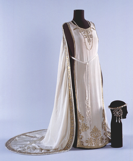 White floor length gown positioned on a black mannequin. The gown features gold silk floral embroidery, and a train rimmed with gold silk detailing. A tie is positioned around the waist and a white necklace sits around the mannequins neck. Positioned on the ground is a head shaped mannequin with a decorative head piece positioned on top.