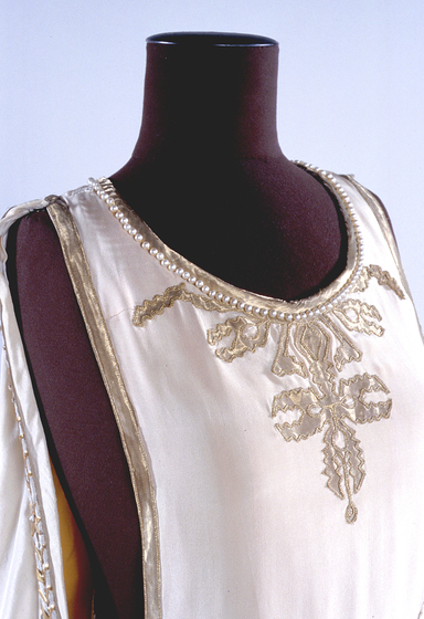 Zoomed in photograph of a white silk gown positioned on a black mannequin. The gown features gold silk embroidered detailing and pearls sewn around the neckline.