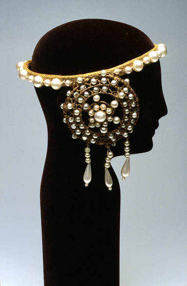 Detailed head piece positioned on a black head mannequin. The gold embroidered head band has pearls of alternating sizes around the rim, and covering the ears is a circular attachment, covered in more pearls and hanging beads.
