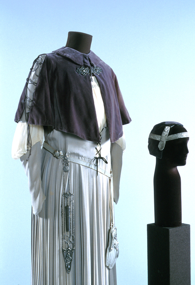 White gown positioned on a black mannequin. Over the white gown is a blue or purple bolero, clasped at the next with a beaded attachment. Hanging from the waist of the gown are silver chains. Next to the mannequin is a head mannequin with a head piece sitting on top.