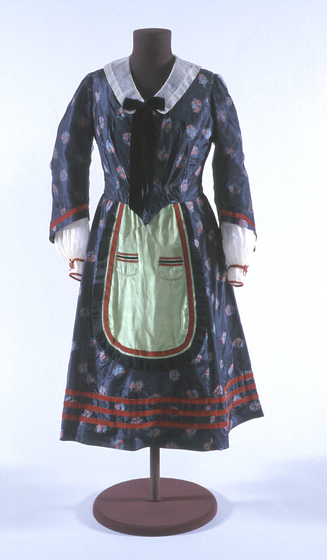 Blue floral dress with full length sleeves positioned on a black mannequin. The dress features an apron type design from the waist down, and a black bow attached to a white collar.