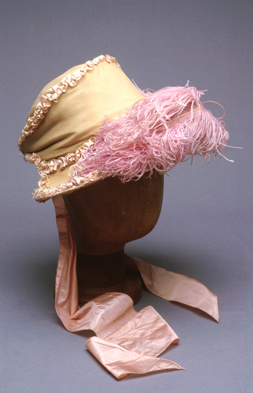 A cream coloured structured bonnet sitting on a wooden head mannequin. The bonnet features pink feathers across the rim and pink silk ribbons hanging off the back.