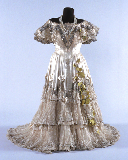 Highly decorated silk dress with full length tiered skirt. Featuring silver and gold sequins, lace, bows and flowers. Pearl necklaces are draped over the mannequins neck.