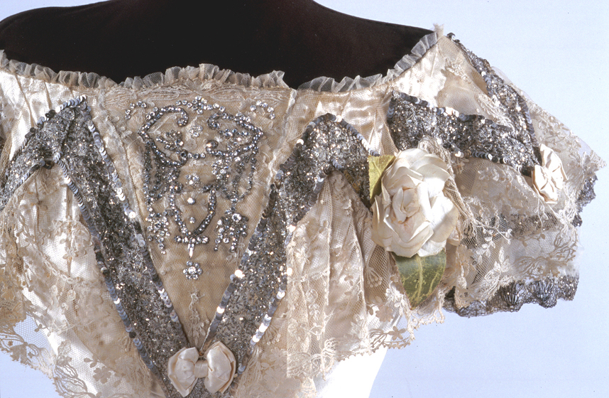 Zoomed in photograph of the detailing of the bodice of a cream colored dress. the bodice features silver sequins, lace, cream bows and flowers and a ruffled silk neckline.