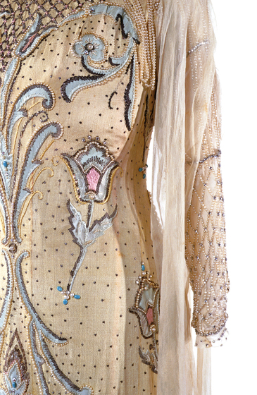 Zoomed in photograph of part of a cream coloured dress, On the bodice of the dress is embroidered flowers in blue and pink. The selves and neckline feature beaded adornments.