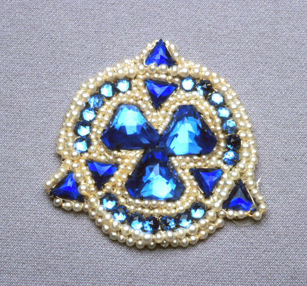 Circular brooch with three triangle points. The brooch is made of small white beads and larger blue gems.