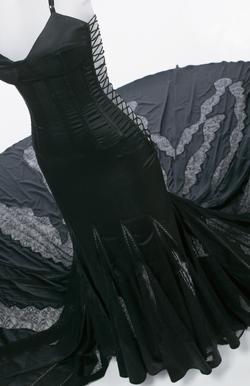 Mannequin wearing black silk corset dress with black lacing at sides and antique lace panels throughout the skirt, which is trailing on the floor.