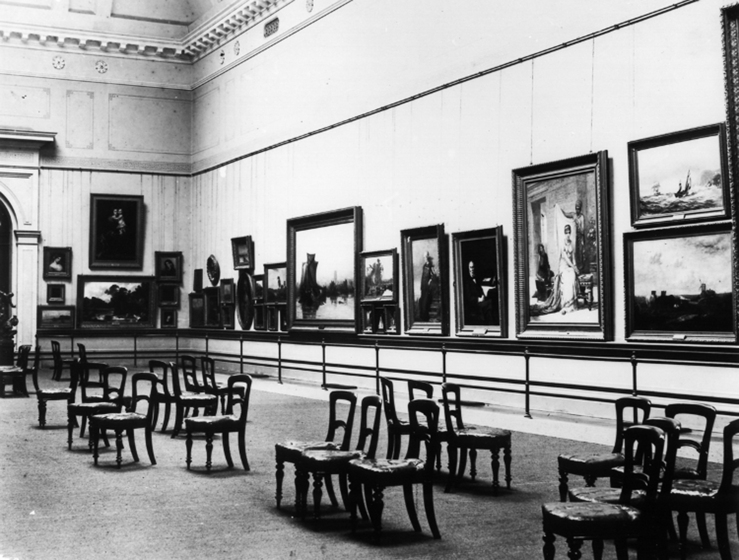 Interior of a gallery with lots of various size framed paintings hanging on the peripheral walls. In the middle, groups of chairs face the paintings.
