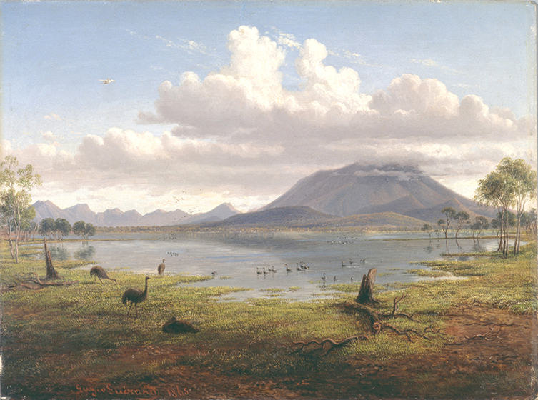 A landscape scene of a large lake or wetland, with sparse open greenery on either side, and on the far bank a large mountain rising up, it's top obscured by clouds. In the foreground emu's graze on the grass, and birdlife can be seen swimming in the water.