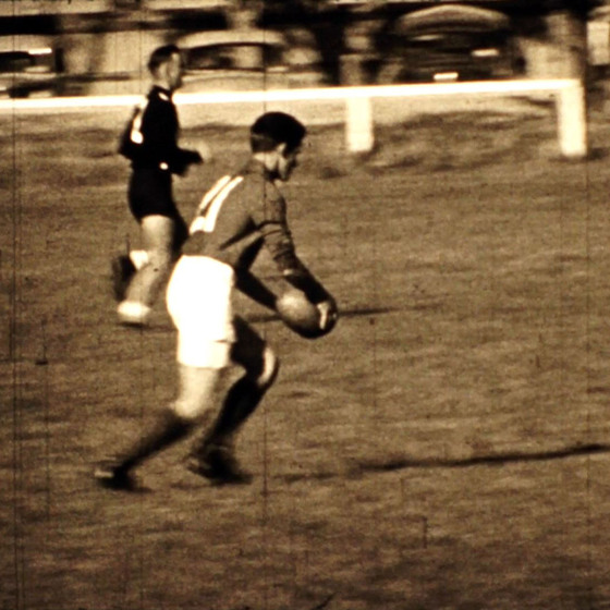 Black and white image of a man wearing a long sleeved football jersey, lining up to kick a football. Another player runs slightly behind him. 