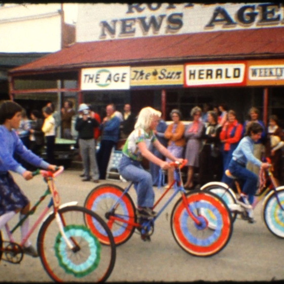 A group of children ride bicycles with decorated wheels. Behind them, people line the street, under shop awnings, to watch the cyclists.