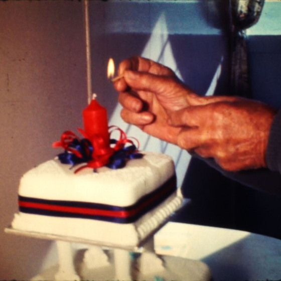 A set of older persons hands light a red candle that is positioned in the centre of a white square cake, wrapped in a blue and red ribbon.