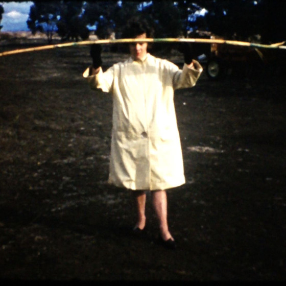 A woman in a white smock and black gloves holds up a long length of slightly bent cane in front of her face.