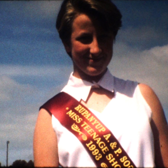 A woman with a short cropped haircut and white sleeveless blouse, smiles at the camera. She is wearing a red sash indicating a place in the Rupanyup Show Miss Teenage pageant.