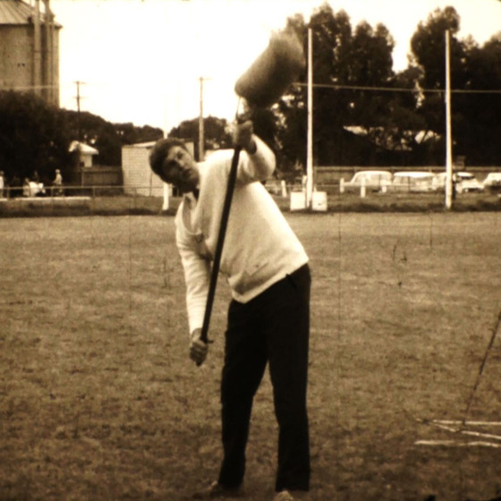 A man stands in the middle of a football pitch, he is holding a pole slightly up in the air, and attached to the end is a sheaf. It appears as though he is trying to throw the sheaf in the air.