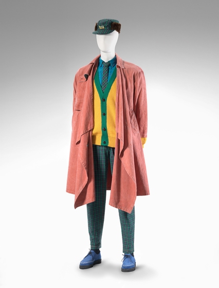 A mannequin dressed in green tartarn pants, a yellow and green cardigan, a faded red canvas coat and a trapper hat. On his feet he has blue suede shoes.