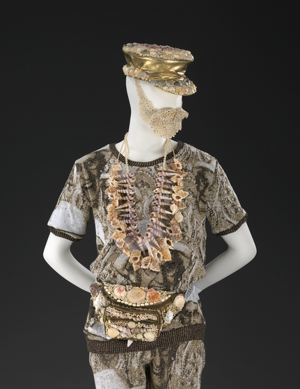A mannequin dressed in tshirt and shorts made of wool, cotton, leather and metal. Around his neck is a large shell necklace, and around the waist a bumbag encrusted with shells and pearls. He is wearing a gold hat, also encrusted with shells on the top and the peak.