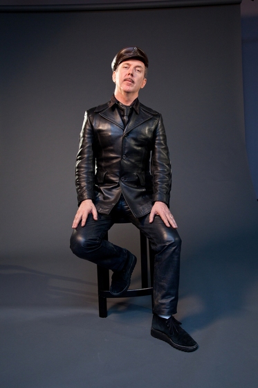 Man sits on a stool dressed in leather pants, jacket and cap