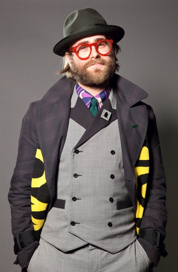 Man dressed in grey checkered two piece suit with matching waistcoat. He wears a blue tartan overcoat, a green fedora styled hat and think rimmed red glasses.