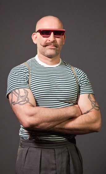 Man dressed in grey pants, green striped t-shirt and suspenders. He has large anchor tattoos on his arms and is wearing sunglasses.