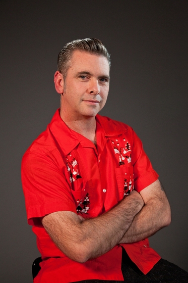 Man dressed in red collared shirt with embroidered segments all over the front. His arms are crossed over his front and his hair is slicked back.