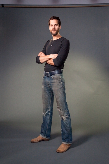 Man stands with his arms crossed across his body, wearing blue jeans, a black belt and a long sleeve black skivvy.