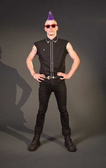 Man stands with his hands on his hips, wearing black combat boots, black jeans, a black button up sleeveless shirt and red sunglasses. He has a bright purple mohawk.