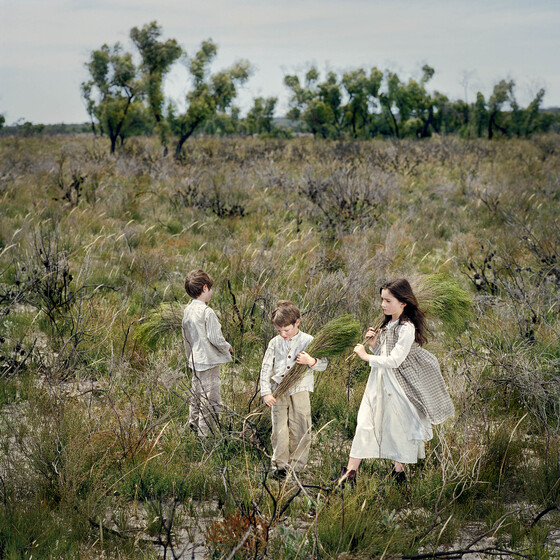 Three young children walking through green scrub. They are holding sheaths of grass and dressed in clothing from the early 19th Century.