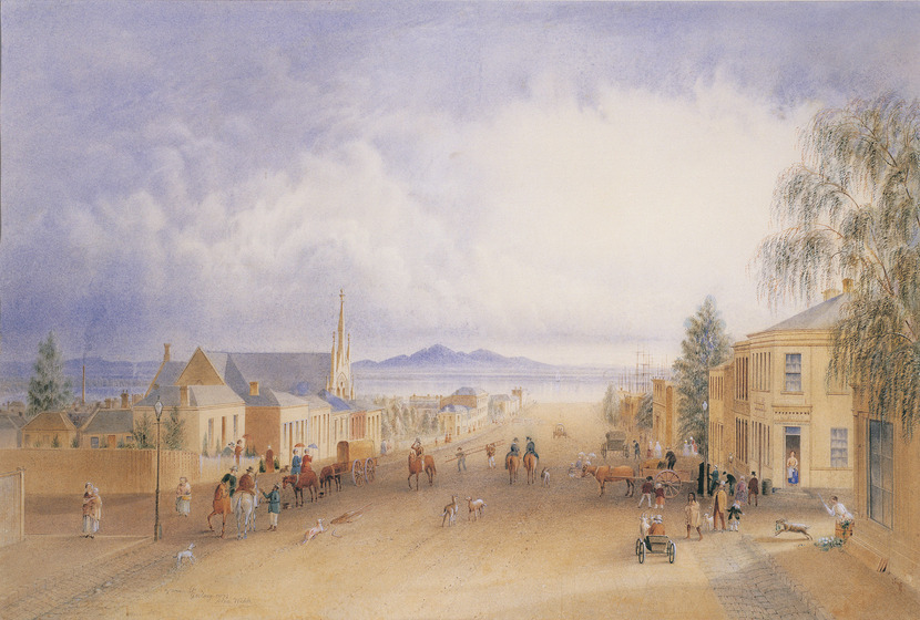 A streetscape of a group of sandstone buildings on either side of a dirt road, including single story, double story and a church steeple. In the street are people walking, in horse and carts and on horses. Dogs and sheep run through the street. In the background is a large lake and hills raising up on the opposite shore.
