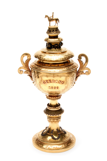 A gold trophy cup, with a lid and on top a small statue of a person riding a horse.