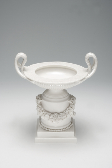 A white porcelain plinth, surrounded by a garland of flowers and beading around the base. On top of the plinth is a white dish with folded over handles on either side.