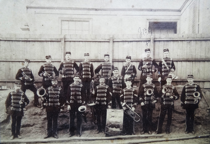 Black and white group photograph of band in matching uniform, including braided coats and hats. All of the men are holding some type of brass instrument, except for one man standing in front of a large drum.