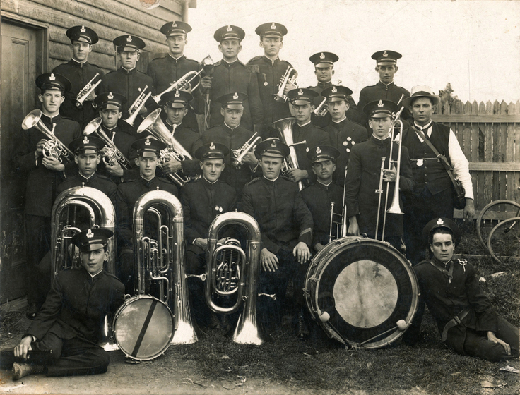 Group of man standing in three rows, all wearing matching black uniforms and peaked caps. Each of the men is holding some type of brass instrument, except for two men sitting on the ground besides two large drums.
