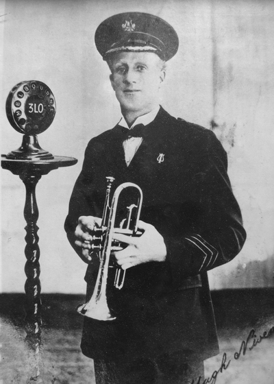 Black and white photograph of a man standing in a black suit, peaked hat and bow tie. He is holding a cornet and next to a wooden table with a circular speaker on top.