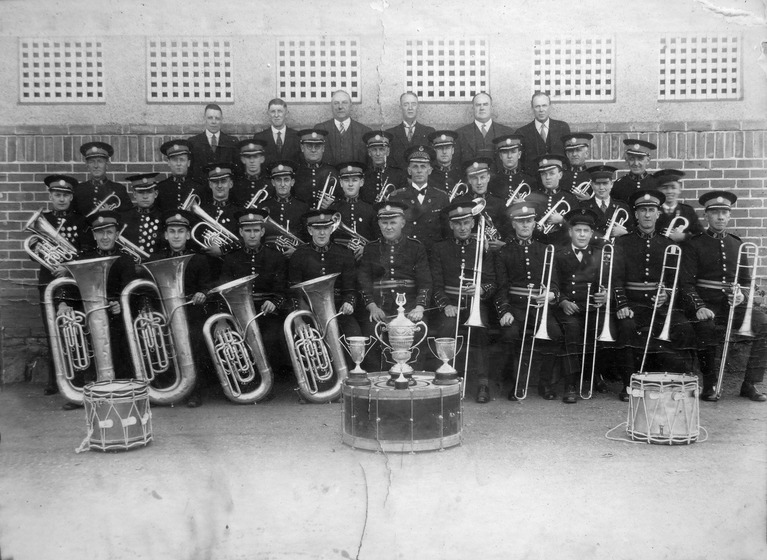 Four rows of men, three sitting in matching uniforms and hats, and the back row standing in suits and ties. The three rows of men in uniform are holding brass instruments, and two drums sit either side of the group on the ground. In the centre on the ground is a drum turned on its side, with four trophies presented on top.
