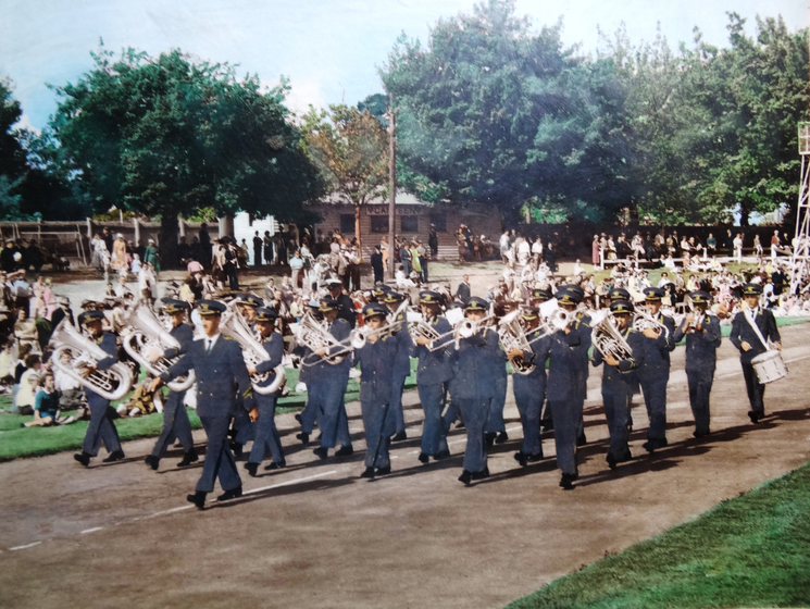 A marching band, all in matching uniform, walk down the middle of a road. On the grass verge on either side, people sit and watch the band.