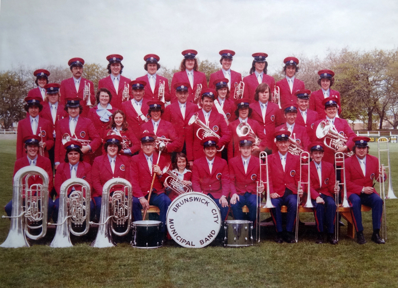 Group photograph of band, all in matching red uniforms and hats, in four rows. The front row is sitting, with the other three rows standing behind each other. All band members are holding some type of brass instrument.