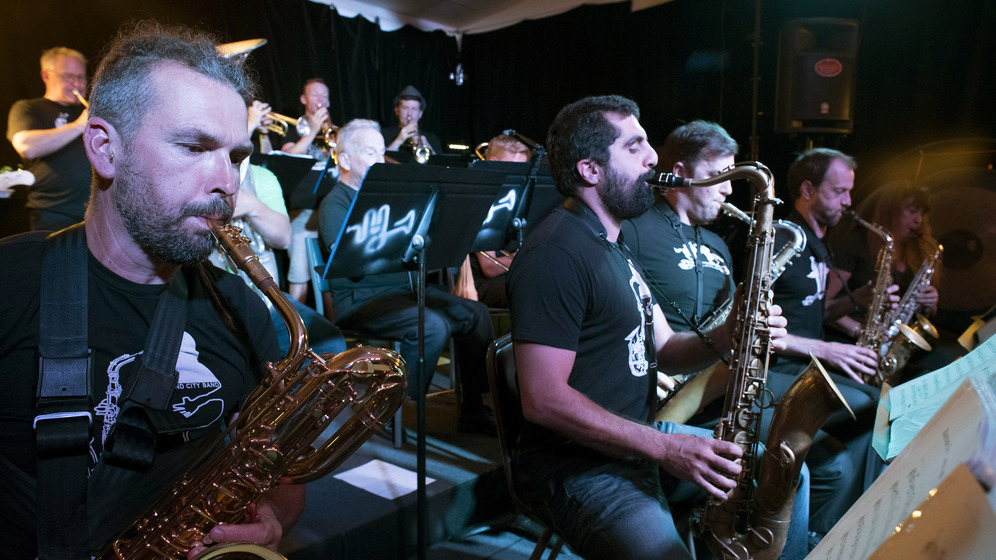 A band sitting on a stage, three rows lined behind each other. The focus is on the front row, all men playing saxophones and reading music on music stands.
