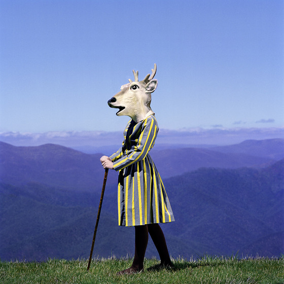 A human figure stands on a grassy patch with rolling blue hills stretching out behind them into the horizon. The figure is dressed in a striped coat and holding a staff, and where the head should be is a deer's head.