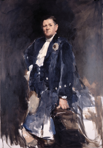 A middle aged man dressed in a blue suit and white shirt, casually leans one hand on a bag, and the other is planted on his hip.