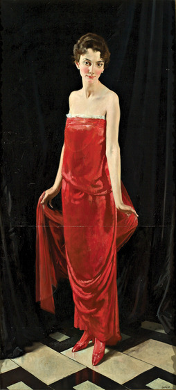 A woman stands in a floor length strapless red dress. She is wearing pointed red heels, and holding in both hands a silk train that falls towards the floor.