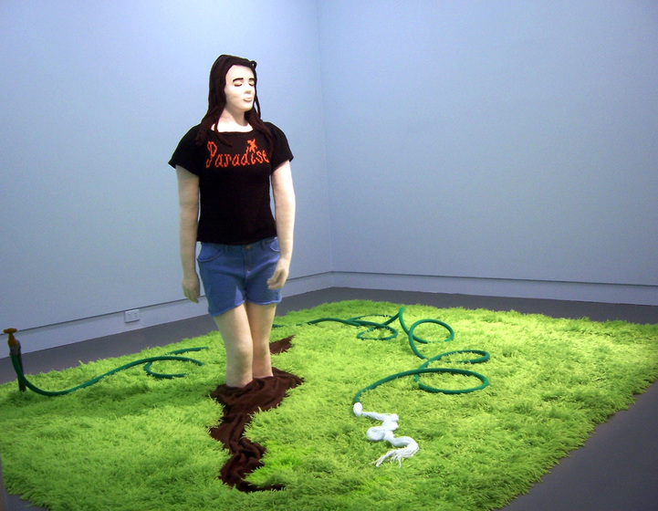A knitted scene of a woman with her feet stuck in a puddle of mud, surrounded by a grass patch. A hose with water spurting out of it lies on the grass. The woman is wearing blue shorts and a black t-shirt with a slogan on it.