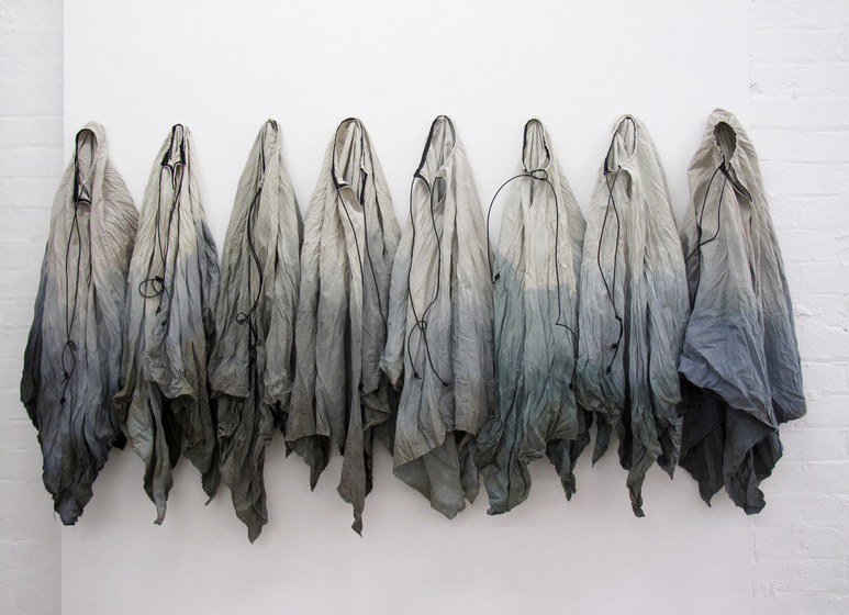 Eight pieces of cloth, hombre from grey blue, hanging on individual hooks on a white wall.
