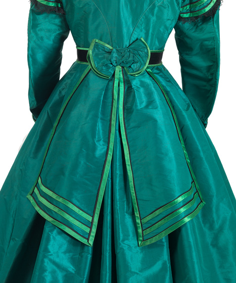 The back of a vivid emerald green full length silk dress. The focus is on a large bow with trailing ends, halfway down the skirt. The edges are sealed with a different shade of green silk