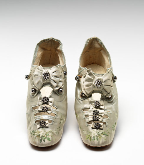 A pair of gold silk closed toe shoes, with a decorative bow clasp, and smaller bows down the front of the toe. The very tip of the shoe is embroidered with green leaves.