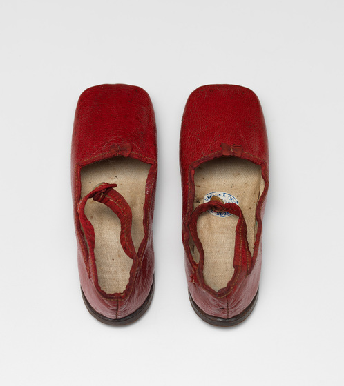 An aerial photograph of a pair of red leather shoes and cream insoles. They have a red ankle strap and a mother of pearl button on the front.