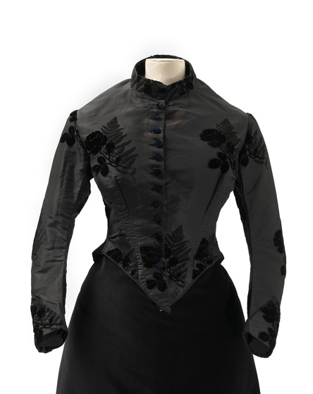 Black silk bodice on a mannequin. The bodice has black embroidered floral emblems over the body and arms, and a button down front with black buttons.