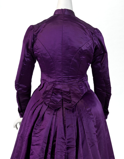 A full length silk dress, with a high neck collar and full length sleeves. There is a small bustle at the back of the dress, indicating the separation between the skirt and top.
