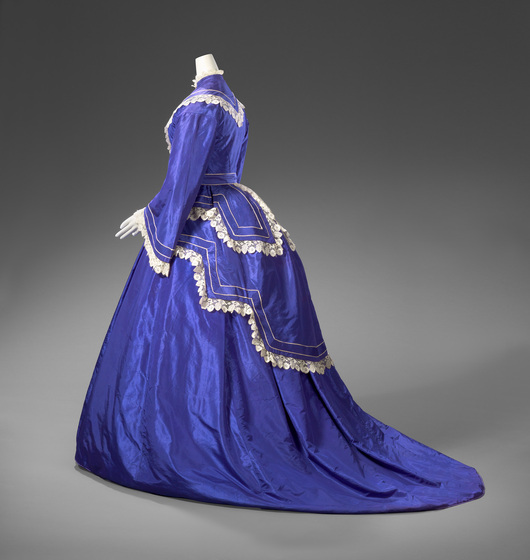 A mannequin wearing a blue floor length silk gown with a decorative white trimmed bustle, neckline and billowing sleeves.