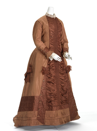 A mannequin wearing a brown floor length gown with a darker brown ruffle paneling down the front, and around the bottom of the dress. The sleeves have buttons from the wrists to almost the elbows, and fringing around the wrists.